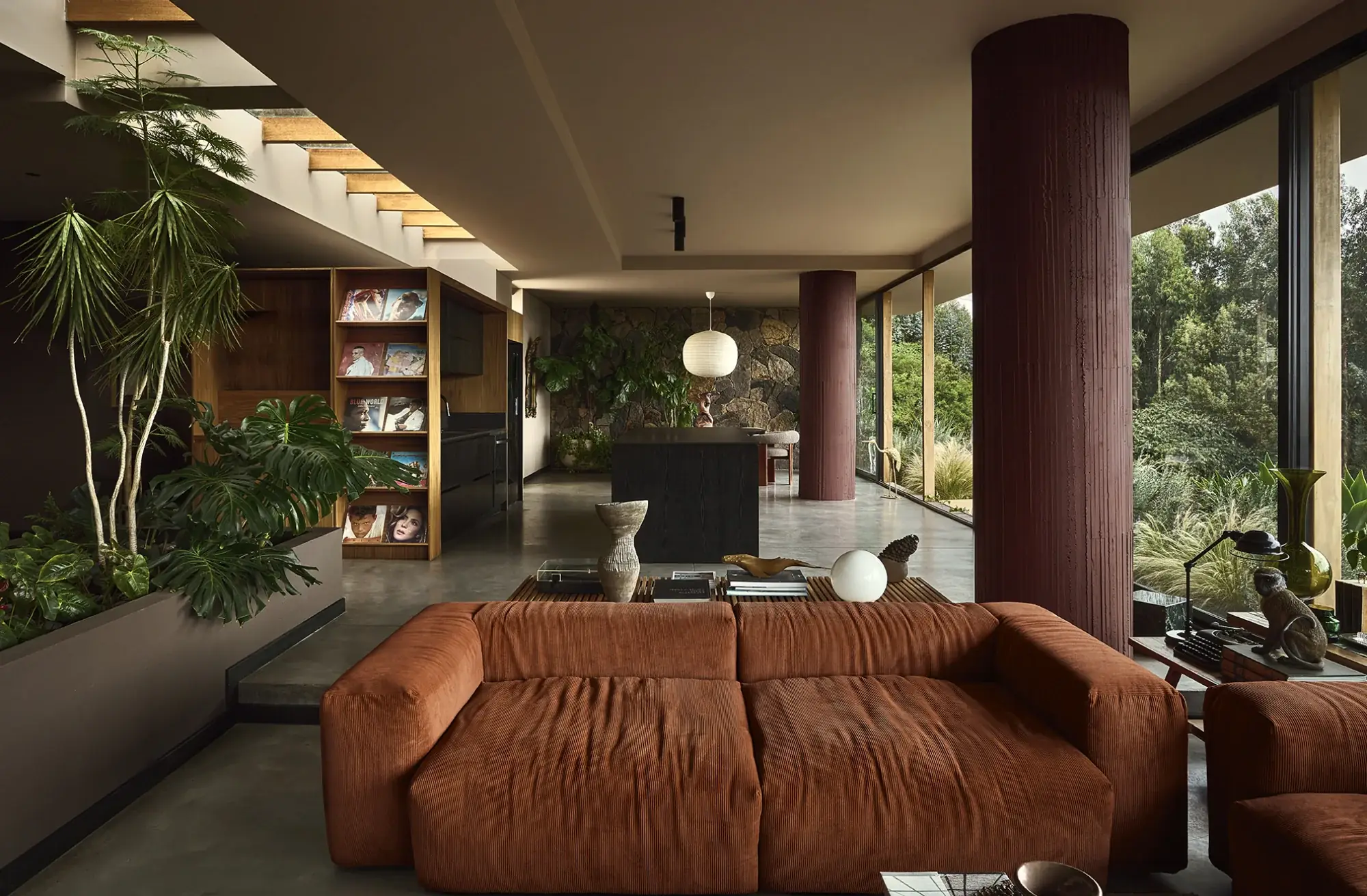 Living residencial con columnas cilíndricas en tono terracota, biblioteca abierta en madera y sofá en pana terracota junto a ventanales
