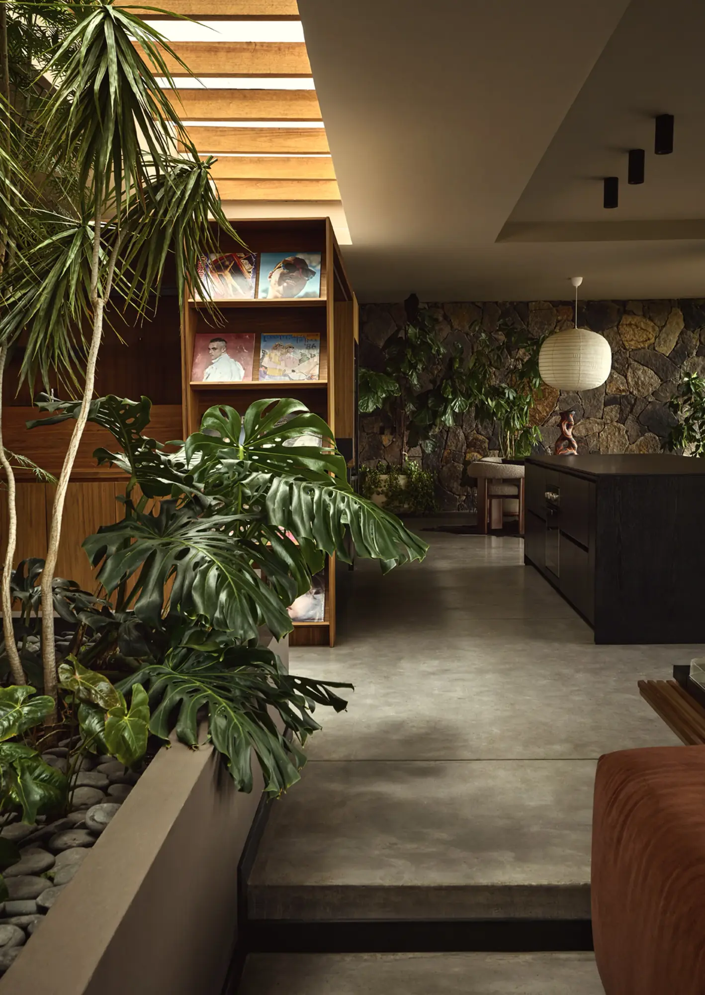 Pasillo residencial con tragaluz de madera, biblioteca artesanal y plantas tropicales junto a una cocina en tonos oscuros