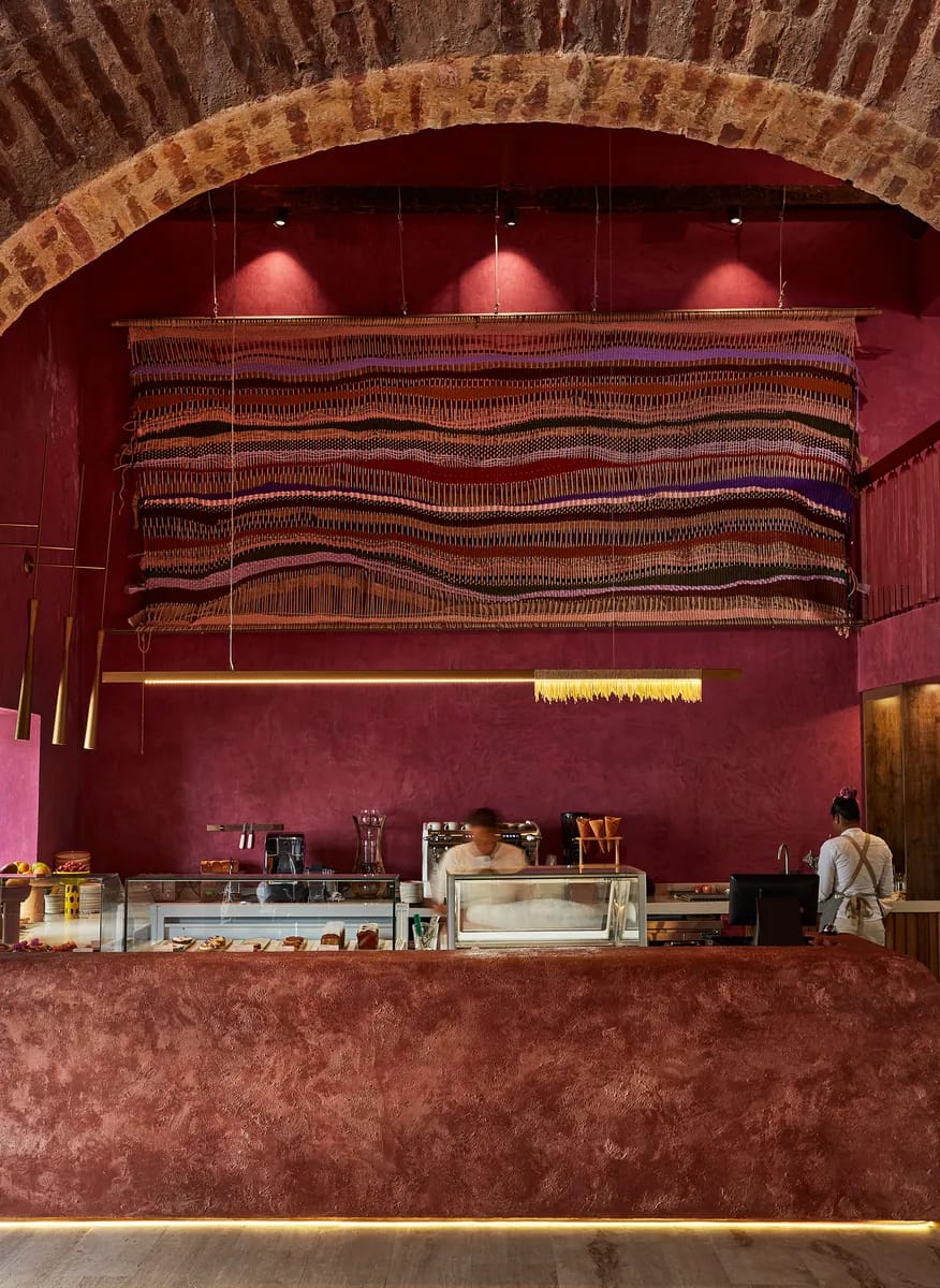 Barra de bar en tono burdeos bajo arcos de piedra, con lámparas colgantes de mimbre en el salón de Nia Bakery