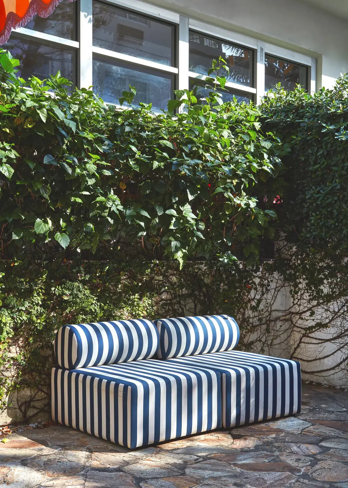 Sofá exterior tapizado a rayas azules y blancas junto a un seto verde bajo una ventana