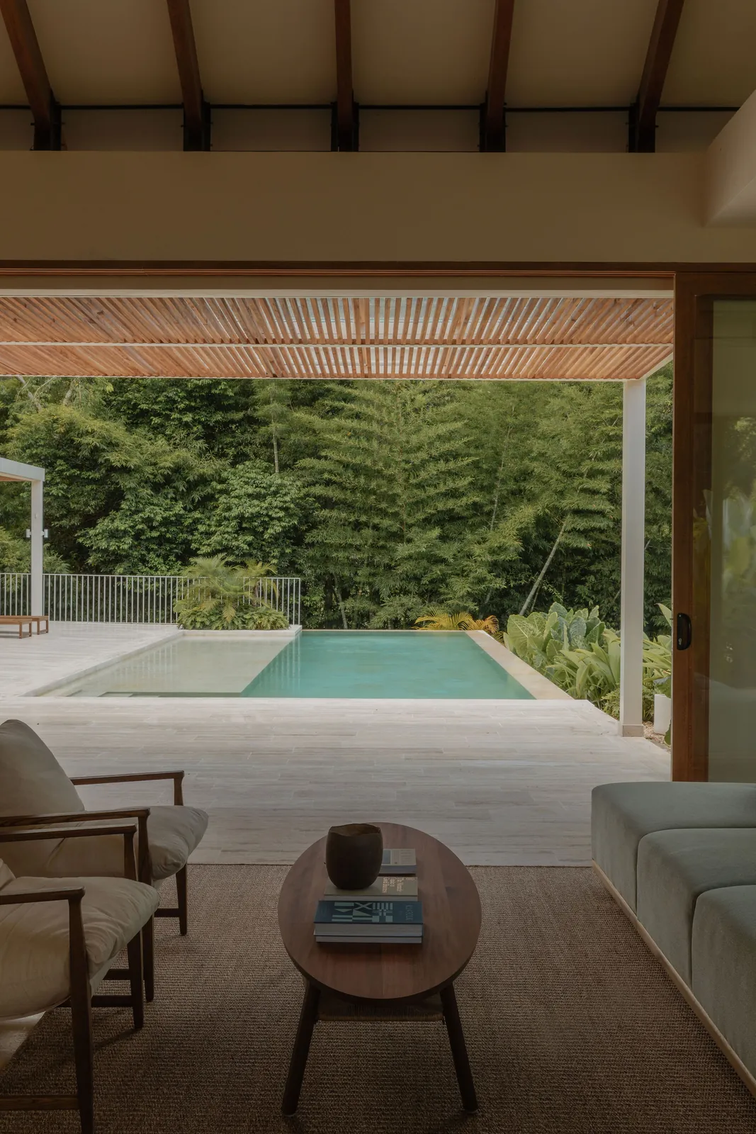 Salón abierto a la piscina y al bosque, con sofá gris, sillones y techo en listones de madera
