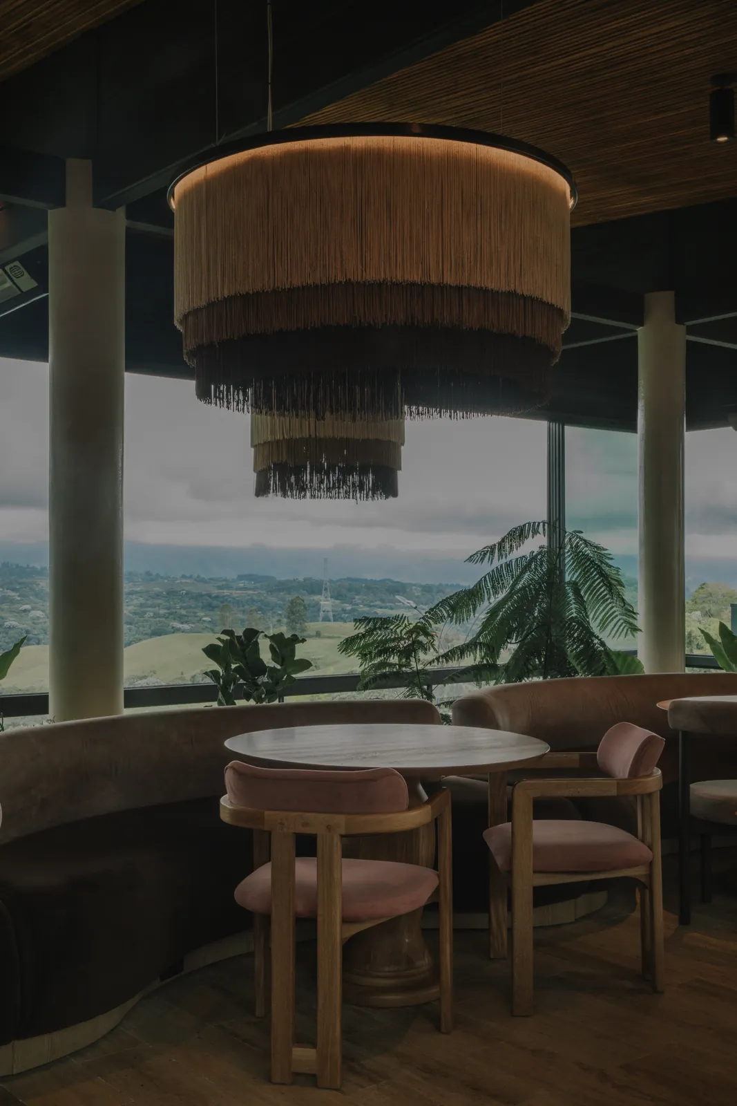Mesa redonda con sillas en terciopelo rosa bajo una lámpara de fleco tejida, con vista panorámica al valle