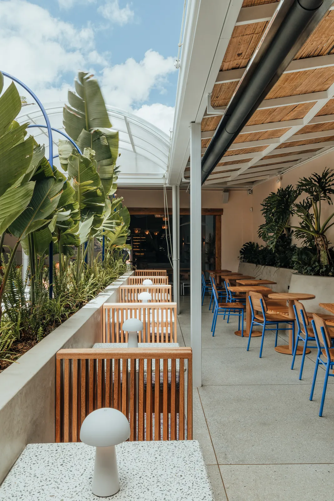 Terraza con mesas de madera y sillas azules bajo un techo de listones junto a un ventanal curvo