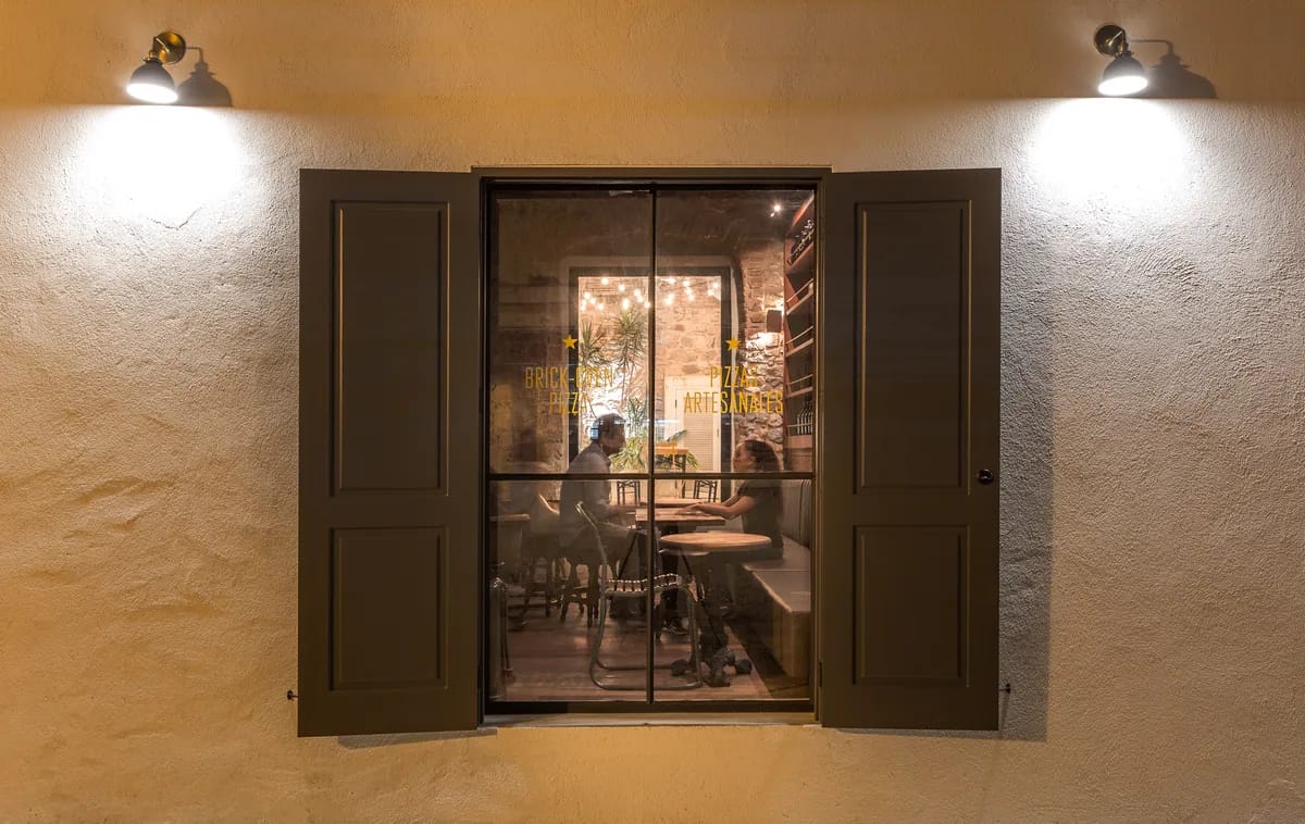 Ventana con contraventanas verdes y letrero de pizzas artesanales sobre muro color mostaza