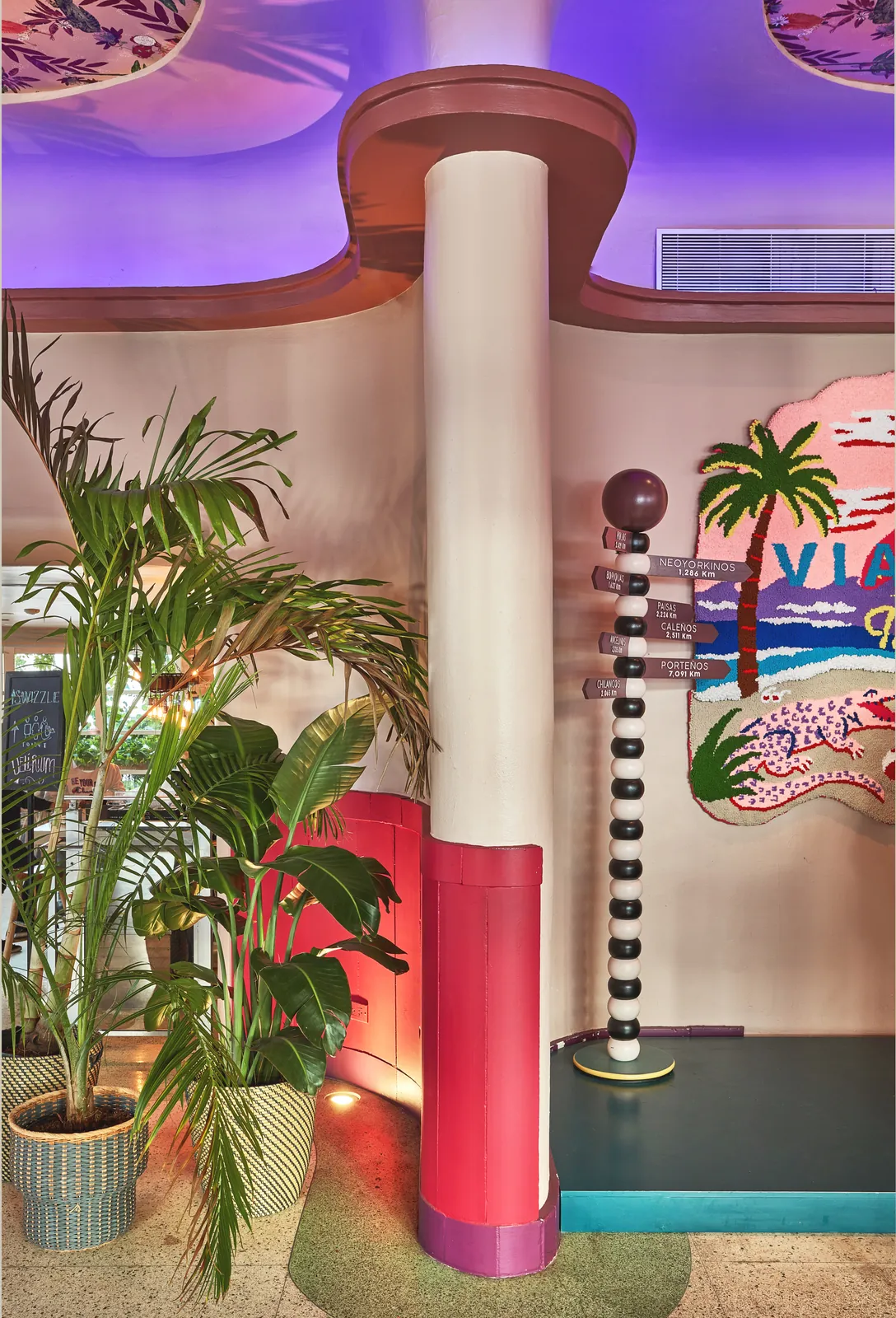 Columna decorativa rosa y roja junto a un tótem señalético con distancias a ciudades, sobre un mural tejido
