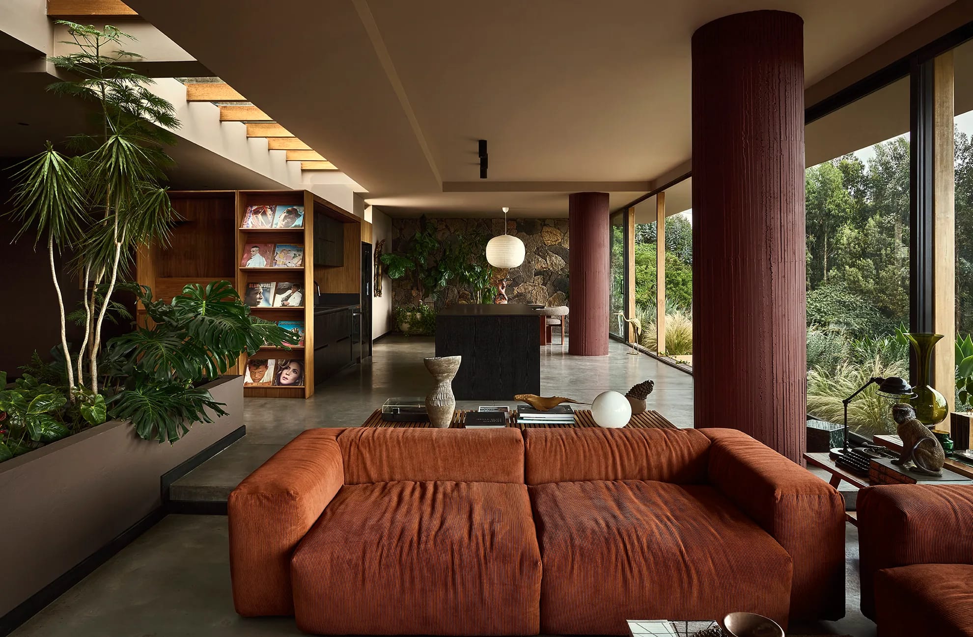 Sala de estar con sofá en pana terracota, columnas revestidas en tono burdeos y ventanales de piso a techo con vista al bosque