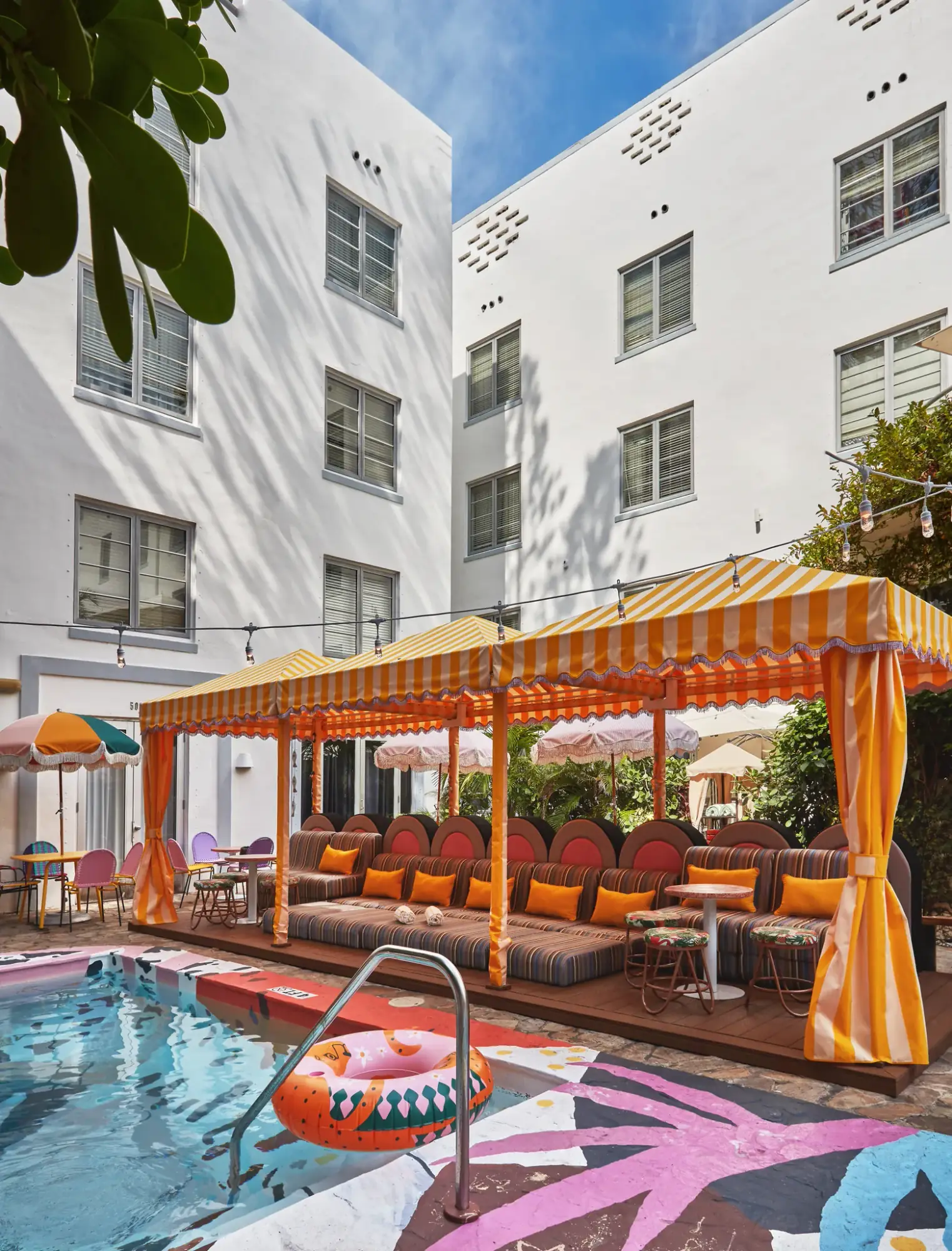 Piscina de Viajero Hostels Miami con cabaña a rayas naranjas, banca corrida tapizada y edificios art déco al fondo