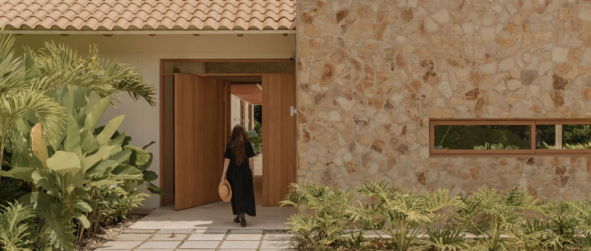 Mujer caminando hacia una puerta pivotante de madera en la entrada de una casa con techo de teja de barro y muro en piedra