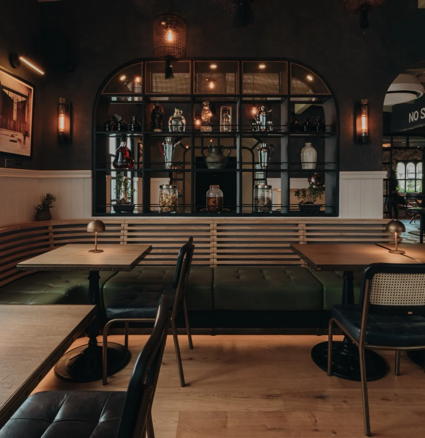 Salón de restaurante con banca tapizada en cuero verde, mesas de madera y sillas de rejilla frente a una barra iluminada