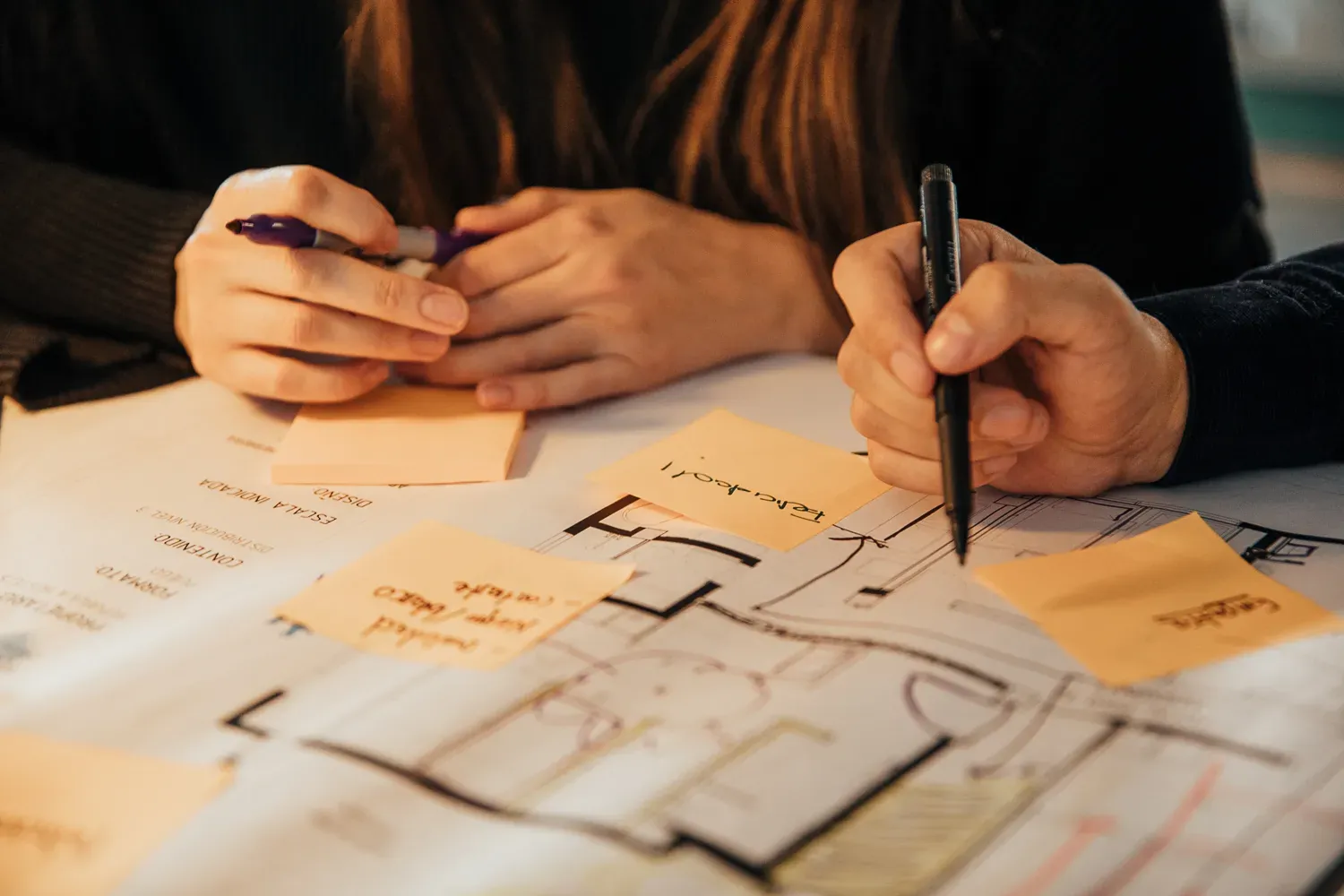 Manos anotando notas adhesivas sobre un plano arquitectónico durante el estudio de distribución espacial